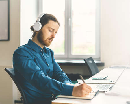 Side view of focused male office worker dressed in casual clothes in headphones watching online webinar on laptop and taking notes of important information in notebook, working on modern officeの写真素材