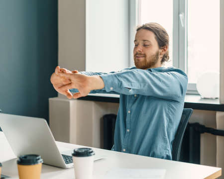 Young relaxed man office worker in casual clothes stretching arms after long work on laptop and looking at computer screen with smile, male employee resting relaxing in office during working dayの写真素材