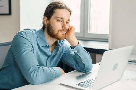 Young unhappy man office worker feeling bored at work, looking at laptop with demotivated face expression while sitting at workplace in office, distracted male worker feeling tired of monotonous jobの写真素材