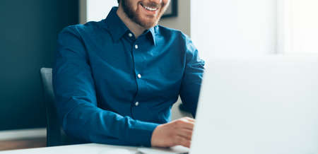 Faceless happy young male office employee in shirt sitting at desk in front of laptop computer with...