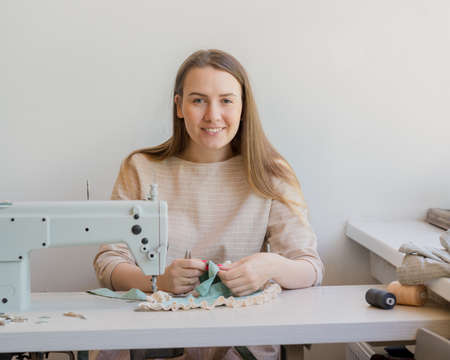Young happy seamstress sewing clothes at her own atelier, smiling at camera. Female atelier owner...