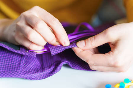 Sewing process. Cropped shot of woman seamstress holding needle while working on new item purple corset at her workspace at home or atelier, close up shot. People and creative hobbies conceptの写真素材