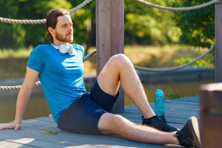 Young relaxed sportive man dressed in sportswear relaxing after workout outdoors in nature,...