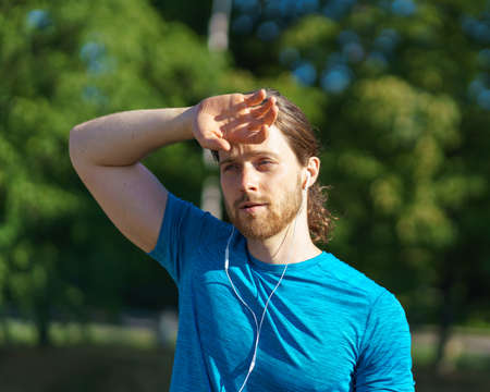 Tired exhausted young sportive man keeping hand on head and resting after hard sports training in...