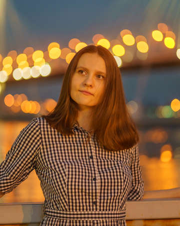 Portrait of young woman on blurred lights of bridge at night or evening in city in summer, beautiful female tourist with long hair looking to urban view, verticalの写真素材