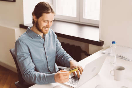 Happy young man sitting at desk in office with credit card and shopping online on laptop, joyful male buyer making payment on computer online, using internet banking service systemの写真素材
