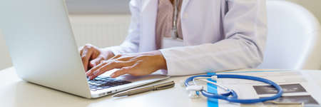 Cropped photo of female doctor in white uniform working on laptop computer in hospital, sitting at...