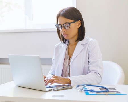 Focused african american female doctor in eyeglasses using laptop while sitting at workplace in medical clinic, young afro woman physician dressed in white coat working on computer in hospitalの写真素材