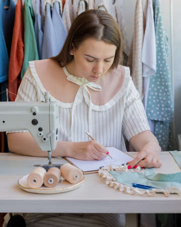 Vertical shot of young woman fashion designer sitting at her workplace with sewing machine in studio and working on new dress collection, drawing clothes sketch. Female seamstressの写真素材