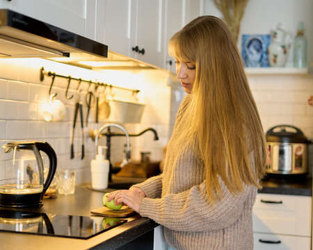 Blond female cutting green apple on cutting board in kitchen. Preparation for cooking recipe. Vegetarian snack. Healthy eating habits. Fruit for breakfast. Vitamin and diet food. Home interiorの写真素材