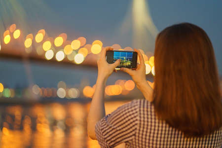 Unrecognizable woman taking photo of beautiful blurred lights of bridge at night or evening in city...