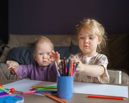 Adorable little girl with golden hair and cute baby boy drawing with colored pencils while sitting at table, older sister playing with small brother while enjoying leisure time together at homeの写真素材