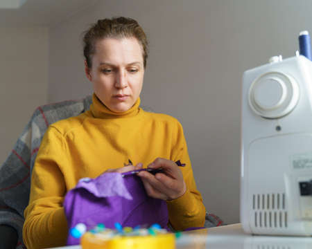 Seamstress concentrated at work. Focused woman in orange sweater working on new clothing item, sewing purple fabric while sitting at her workplace in atelier or home. Female tailor holding textileの写真素材
