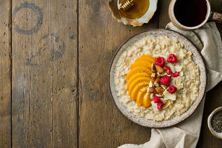 Oatmeal porridge with sliced mango, fresh raspberries, chopped almond nuts, coconut chips, chia seeds, honey and tea. Traditional breakfast served in ceramic plate on wooden background. Copy spaceの写真素材