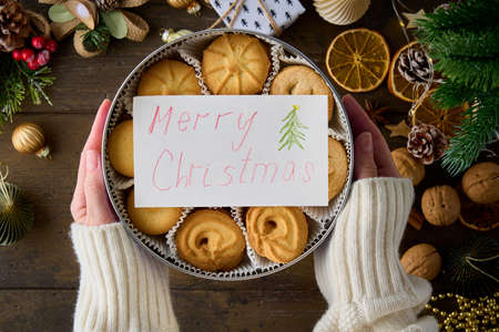 Box with cookies and handwritten note with new year greetings on wooden background. Anonymous female. Christmas decor. Winter bakery. Fir, toy, candle, lemon, berry, paper ball, garlandの写真素材