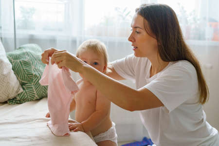 Mommy dressing little girl in dress. Naked baby in diaper standing by sofa and looking at cameraの写真素材