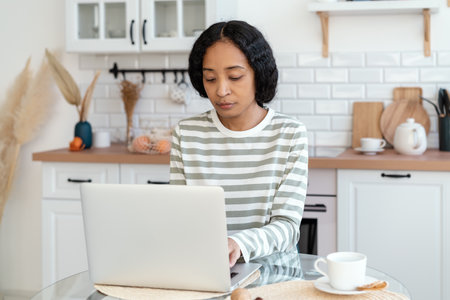 African-american female using laptop while sitting in kitchen. Confident and focused on remote workの写真素材