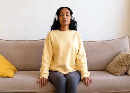 African-american female sitting on couch and listening to music in headphones. Breathing therapyの写真素材