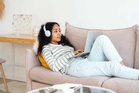 African-american female in headset chatting online on laptop while lying on sofa in living roomの写真素材