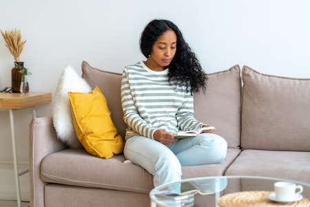 African-american female reading book while sitting on sofa in cozy living room. Spending time aloneの写真素材