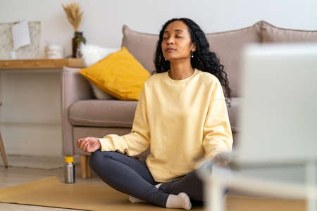 Young african-american female sitting in lotus pose on mat in living room while meditatingの写真素材