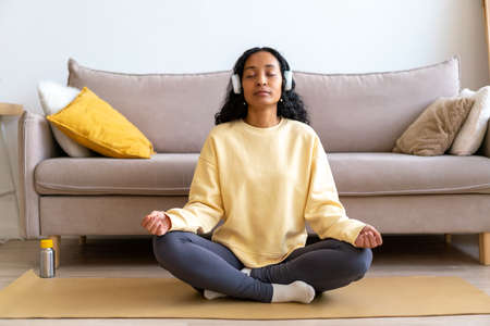 Young african-american female in headphone listening to music while meditating on mat in living roomの写真素材