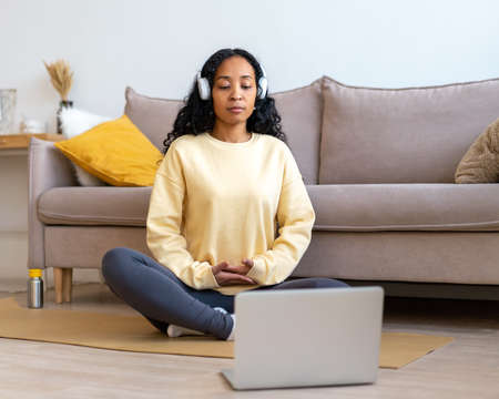 African-american female listening to music in headphones while meditating with laptop at homeの写真素材