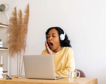 African-american female student yawning while listening to online lecture in headphones on laptopの写真素材