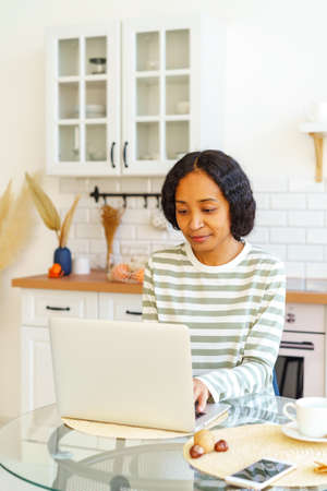 African-American female focused on remote work while sitting in kitchen and using laptopの写真素材