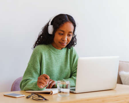 Young African-American female working on laptop in headphones while taking notes in notebookの写真素材