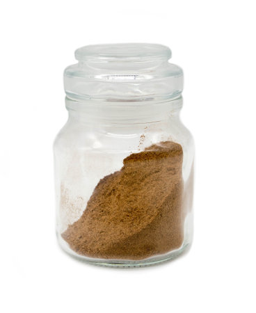 Ground nutmeg in a transparent glass jar on a white background.