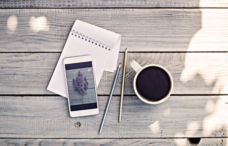 Cup of coffee, notepad, phone with photo of lilac branch on the white wooden tableの写真素材
