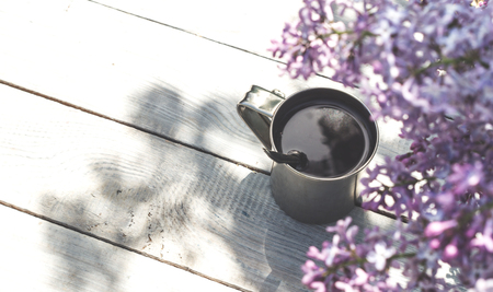 A metal mug with coffee and lilac branches on the wooden tableの写真素材