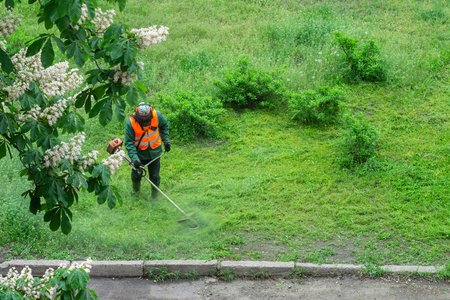 A communal service worker in an orange vest mows grass in a park with a hand lawn mower