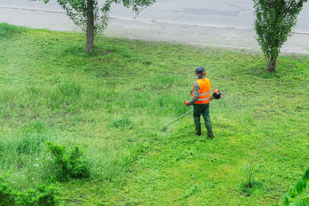 A communal service worker in an orange vest mows grass in a park with a hand lawn mower