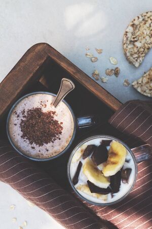 Two glass cups with ingredients banana, chocolate, oatmeal and yogurt and cooked smoothie in a wooden box. Top viewの写真素材