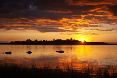 Rising sun, orange, red and deep blue clouds over a quiet lake at sunriseの写真素材
