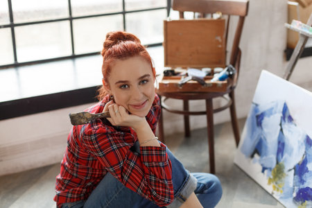 Portrait of a young beautiful redhead female artist painting. Smiling girl sitting on the floor in art studio and looking at the cameraの写真素材