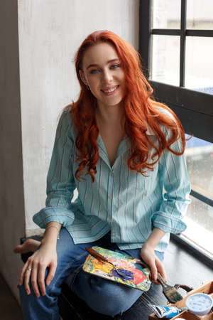Portrait of a young redhead female artist painting. Smiling girl sitting on a windowsill with a palette in her hands and looking at the cameraの写真素材