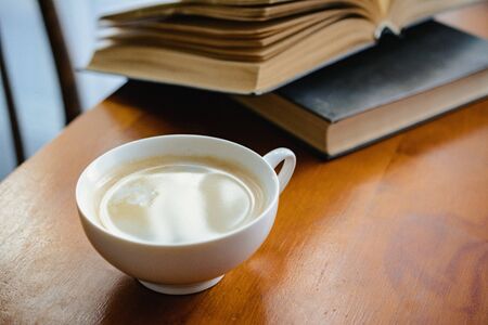 a Cup of coffee with milk on a wooden table with an open book in the background. light from the window. the concept of reading books over a Cup of morning coffeeの写真素材
