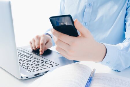 a man in a blue shirt is working on a laptop, holding a smartphone in his hands. Remote work, remote...