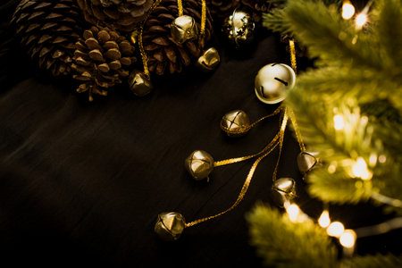 Golden bells on a black background with bright lights of a Christmas garland. Christmas toys lie with pine cones and green fir branches. Bright lights of garlands on the branches of a Christmas tree.の写真素材
