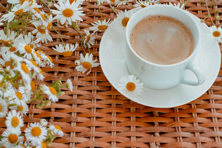 a white cup of coffee with a bouquet of daisies on the wicker table, morning coffee. Cappuccino in a white cup and saucer. horizontal background imageの写真素材