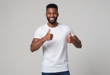 A happy man in a white T-shirt giving two thumbs up, showing a toothy smile. Studio shot with a grey background.の素材