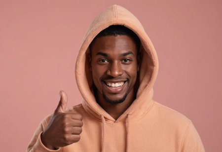 A joyous man in an orange hoodie giving a thumbs up with a warm smile. Pink background.の素材