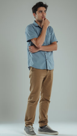 A pensive young man stands against a soft gray backdrop, dressed in a light blue shirt and tan pants. His thoughtful expression and relaxed posture evoke a sense of contemplation and introspection, inviting viewers to ponder the thoughts behind his gazeの素材