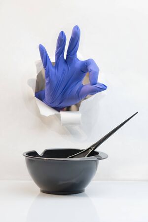 Hand in blue hairdressing gloves through a hole shows gesture all well. A black bowl with a brush is ready for stainingの写真素材