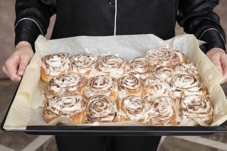 Female hands holding tray of cinnamon rolls or cinnabar, homemade sweet traditional dessert buns with white cream sauceの写真素材