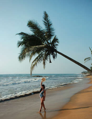 young woman stands under a palm tree on the beach .の写真素材