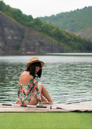 a girl in a swimsuit with sleeves sits on the pier. rocks and river